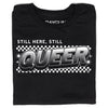 Black, union-made t-shirt featuring the text STILL HERE, STILL QUEER in a bold, comic-style font over a black and white checkerboard background.