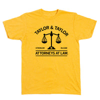 A heather gold shirt with black ink text that reads "Taylor & Taylor: Attorneys At Law" with in image of a scale that has a basketball on either side