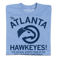 Folded heather light blue t-shirt with navy ink that reads "The ATLANTA HAWKEYES! The Official Sports Team Of The Vast Left-Wing Conspiracy!" With a bird logo in the center.