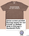 A brown, garment-dyed heavyweight t-shirt that says TOGETHER, WE ARE AMERICA, with a text overlay noting that proceeds support Somos Arte in Chicago’s Humboldt Park.