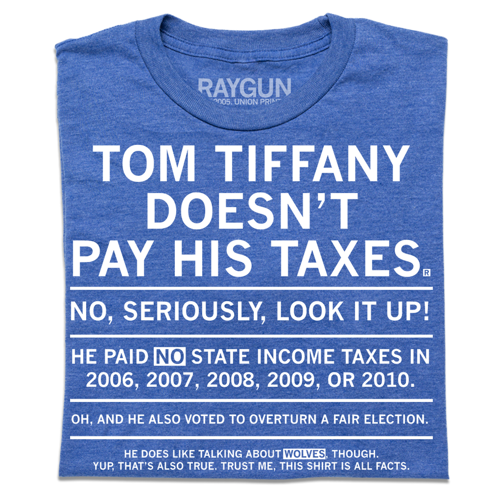 A folded heather royal blue t-shirt with centered white ink text that gets smaller every sentence. It says, "Tom Tiffany Doesn't Pay His Taxes. No, seriously, look it up! He paid NO state income taxes in 2006, 2007, 2008, 2009, or 2010. Oh, and he also voted to overturn a fair election. He does like talking about wolves though. Yup, that's also true. Trust me, this shirt is all facts."