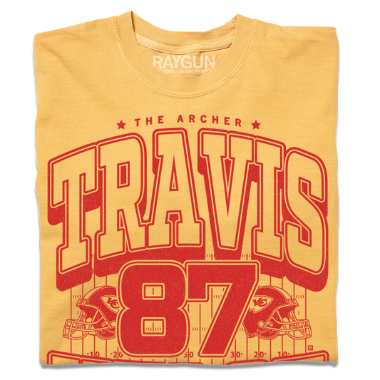Gold shirt with bright red ink that reads "The Archer. Travis Kelce. The Greatest Tight End Of All Time" over an illustration of a football field  with the number 87 and a football helmet on either side.