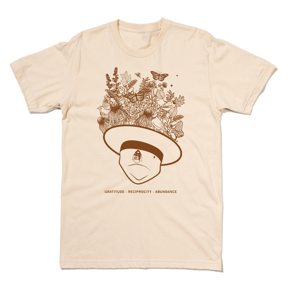 A cream t-shirt with brown ink depicting an upside down park ranger hat with flowers, leaves, birds, and pollinators emerging from it. Text below reads "Gratitude - Reciprocity - Abundance"