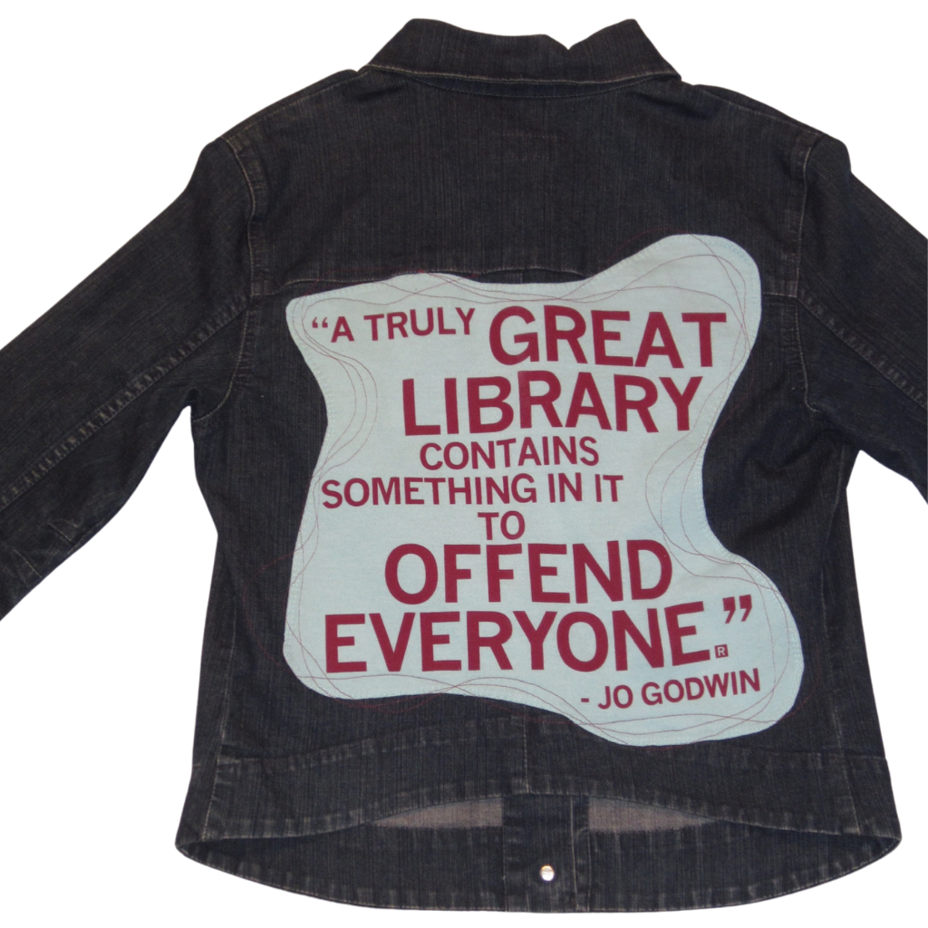 Dark wash denim jacket featuring a light blue graphic on the back with the text, A TRULY GREAT LIBRARY CONTAINS SOMETHING IN IT TO OFFEND EVERYONE. - Jo Godwin.