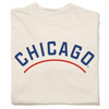 Folded ivory heavyweight t-shirt with "CHICAGO" printed in curved blue font. A red arch is printed below the text, parallel to it.