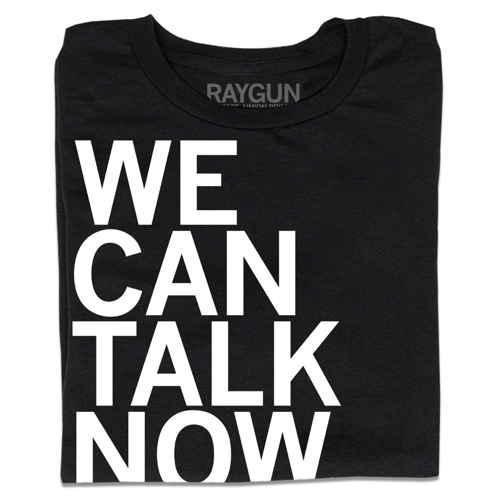 A folded black t-shirt with left-aligned white ink text that reads "We Can Talk Now"