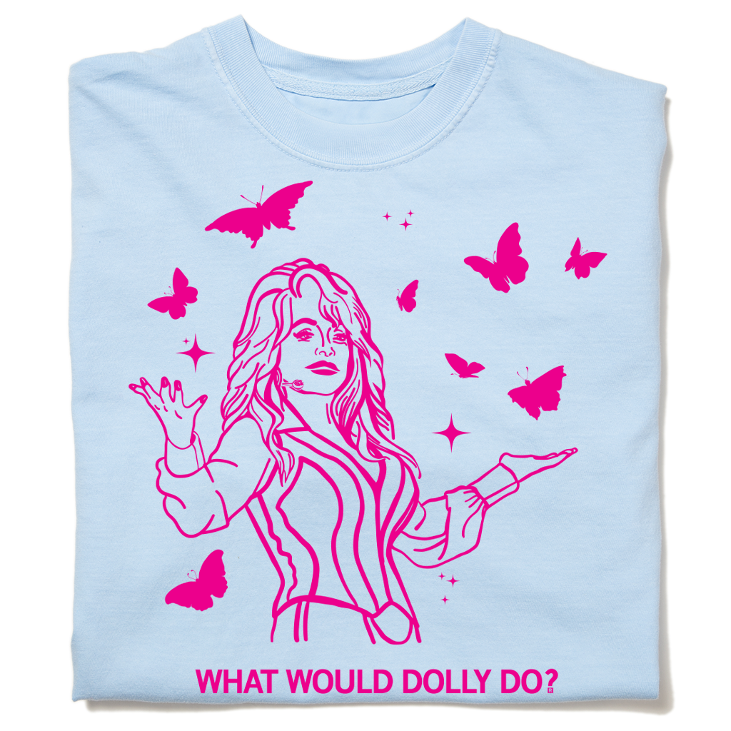 A folded pastel blue heavyweight t-shirt with hot pink ink depicting Dolly Parton with open arms, surrounding by butterflies and sparkles. All caps text below says, "WHAT WOULD DOLLY DO?"