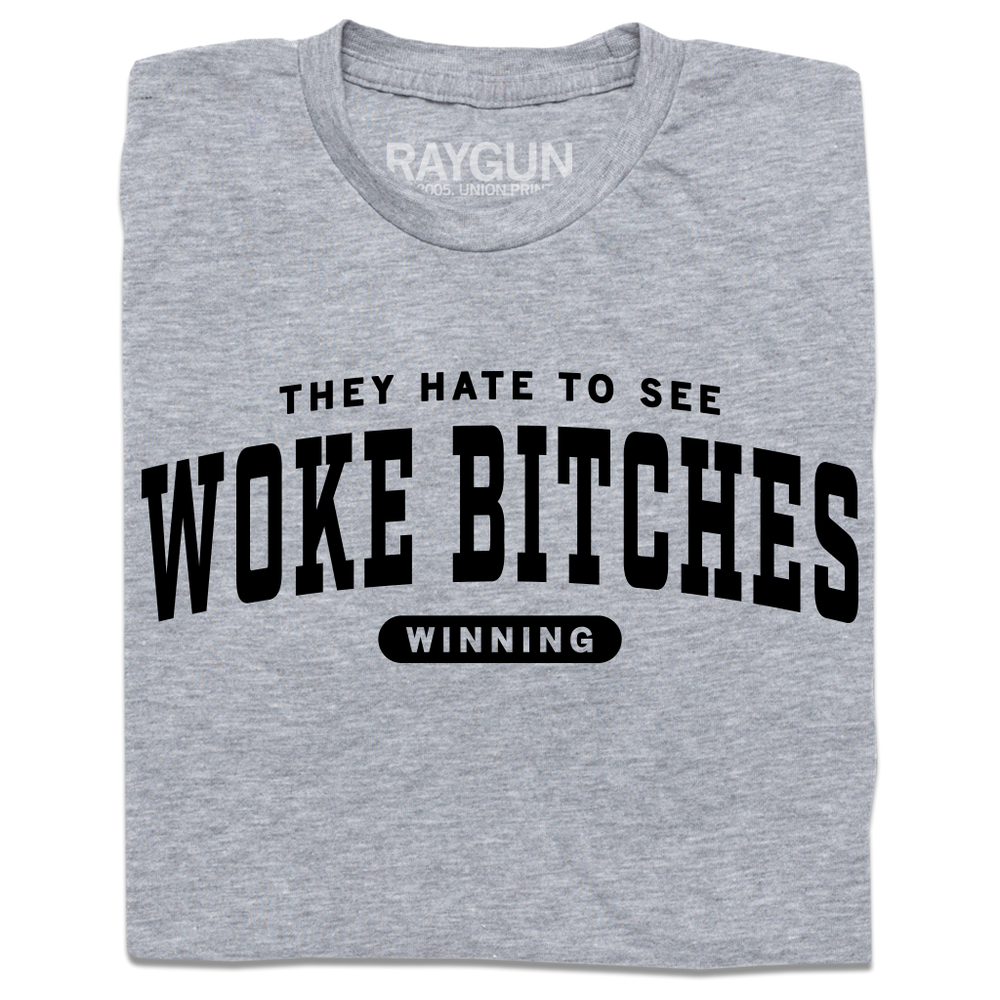 A folded heather grey t-shirt with black ink text that reads "They hate to see woke bitches winning"