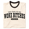 A folded ringer shirt with a cream base, black collar, and black around the sleeves. Centered black ink text says, "A folded heather grey t-shirt with black ink text that reads "They hate to see woke bitches winning"