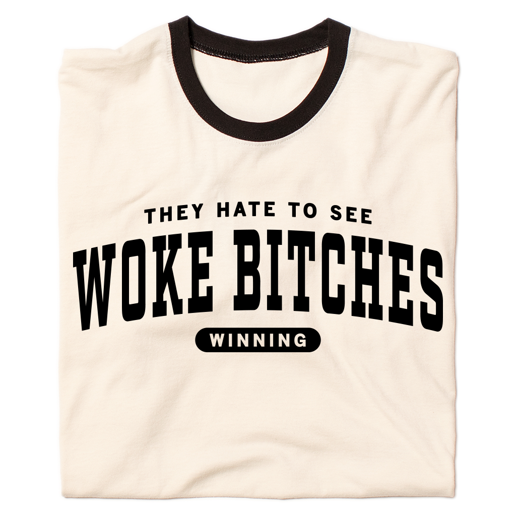 A folded ringer shirt with a cream base, black collar, and black around the sleeves. Centered black ink text says, "A folded heather grey t-shirt with black ink text that reads "They hate to see woke bitches winning"