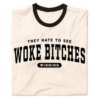 A folded ringer shirt with a cream base, black collar, and black around the sleeves. Centered black ink text says, "A folded heather grey t-shirt with black ink text that reads "They hate to see woke bitches winning"