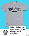 Vintage white ringer t-shirt with black text reading THEY HATE TO SEE WOKE BITCHES WINNING, featuring a cat character in a speech bubble that says, Woke bitches out doing things. Like winning gold medals.