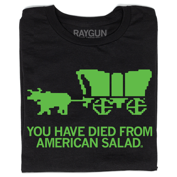 A folded black t-shirt with bright green ink. A pixelated imaged at the top is spoofing The Oregon Trail game and shows and ox pulling a wagon. All caps text below reads 