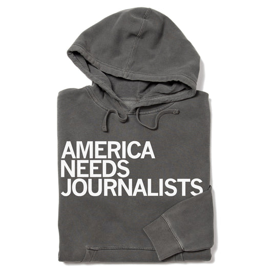 A charcoal gray Pigment Black Pullover Hoodie with the words AMERICA NEEDS JOURNALISTS printed in white on the front.