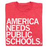 Folded red t-shirt with white text 'AMERICA NEEDS PUBLIC SCHOOLS' on a white background