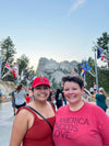 Two women smiling for a photo in front of Mount Rushmore and American flags, with one wearing a heather red T-shirt that reads America Needs Love.