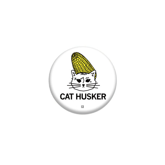 A RAYGUN metal button featuring a line drawing of a cat wearing a corn cob on its head, with the text CAT HUSKER below.