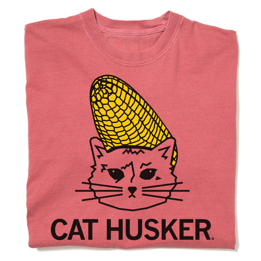 A heavyweight, coral-colored t-shirt featuring a graphic of a cat with an ear of corn on its head and the text CAT HUSKER printed below.