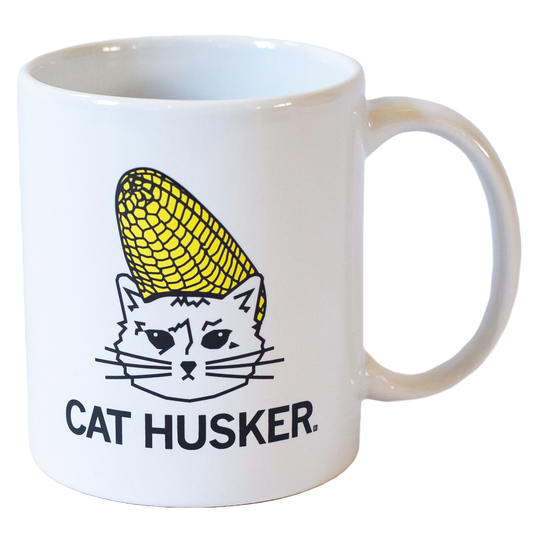 White 11 oz Cat Husker mug featuring a line-art illustration of a cat wearing a corn cob on its head, with the text CAT HUSKER. underneath.