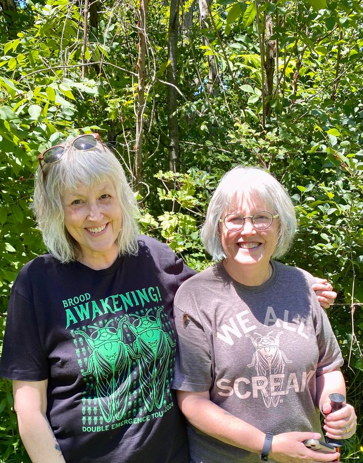 Two smiling women standing in front of green trees, wearing matching cicada-themed RAYGUN T-shirts.