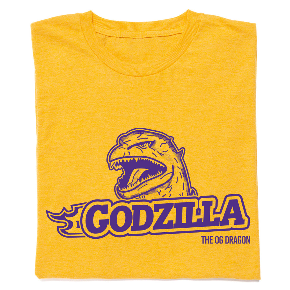 A folded heather gold t-shirt with purple ink that says, "Godzilla The OG Dragon" with Godzilla's head emerging above the text.