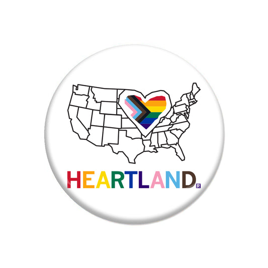 A RAYGUN circular pin featuring an outline of the United States with a Progress Pride flag heart over the center and the word HEARTLAND in multicolored text below.