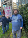 A bearded man in a union-made blue RAYGUN t-shirt that reads I’m going to stop drinking just as soon as I’m named Secretary of Defense holds a sign that says This is what a feminist looks like.