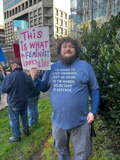 A bearded man in a union-made blue RAYGUN t-shirt that reads I’m going to stop drinking just as soon as I’m named Secretary of Defense holds a sign that says This is what a feminist looks like.