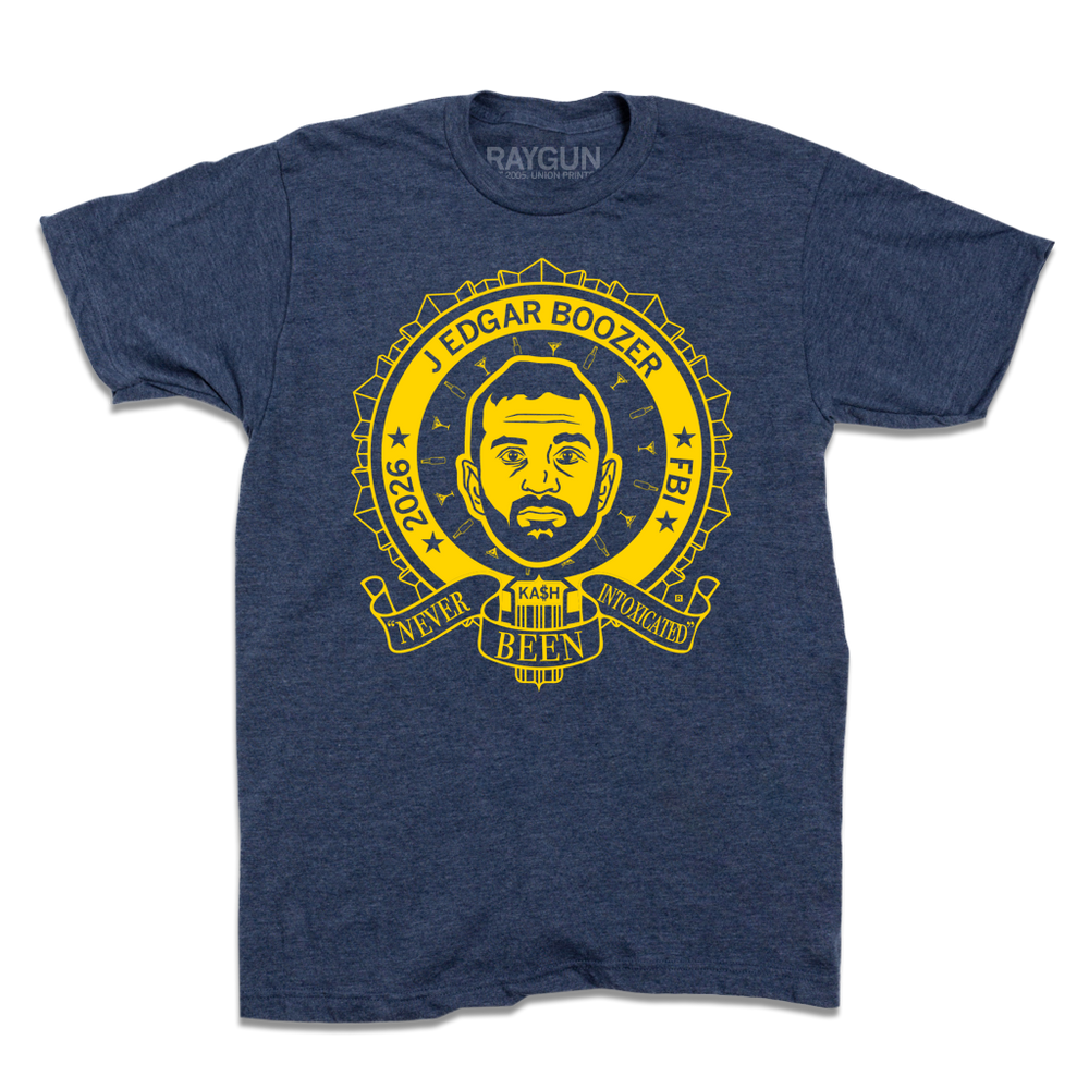 Heather navy t-shirt with bright yellow ink illustrating a ribbon around an image of Kash Patel. Text reads "2026 J Edgar Boozer FBI. Never been intoxicated" with small martini glasses and beer bottles circling his head.