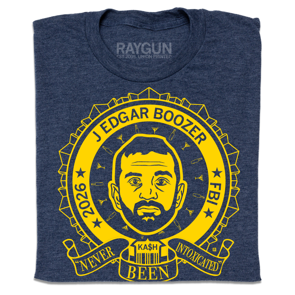 A folded heather navy t-shirt with bright yellow ink illustrating a ribbon around an image of Kash Patel. Text reads "2026 J Edgar Boozer FBI. Never been intoxicated" with small martini glasses and beer bottles circling his head.