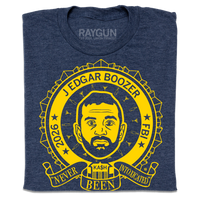A folded heather navy t-shirt with bright yellow ink illustrating a ribbon around an image of Kash Patel. Text reads "2026 J Edgar Boozer FBI. Never been intoxicated" with small martini glasses and beer bottles circling his head.
