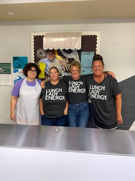 Four cafeteria staff members stand behind a serving counter, three wearing charcoal T-shirts that read Lunch Lady Energy.