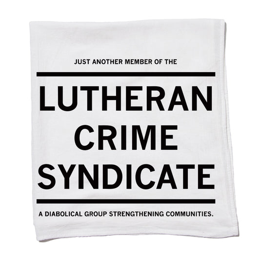White flour sack kitchen towel printed with the text, JUST ANOTHER MEMBER OF THE LUTHERAN CRIME SYNDICATE. A DIABOLICAL GROUP STRENGTHENING COMMUNITIES.