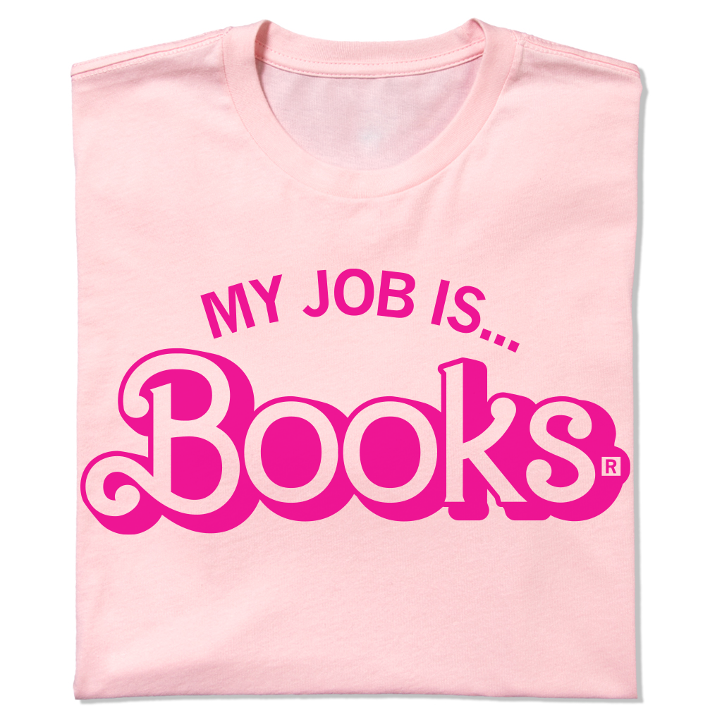 A folded light pink t-shirt with hot pink ink text that says, "My Job Is...Books" with "Books" being in Barbie font.