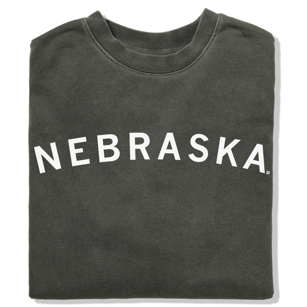 Folded charcoal gray RAYGUN crewneck sweatshirt with the word NEBRASKA printed in white arching across the chest.