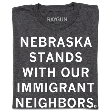 Dark gray union-made t-shirt featuring the text NEBRASKA STANDS WITH OUR IMMIGRANT NEIGHBORS in white block letters.