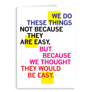 A folded greeting card featuring the text: WE DO THESE THINGS NOT BECAUSE THEY ARE EASY, BUT BECAUSE WE THOUGHT THEY WOULD BE EASY.