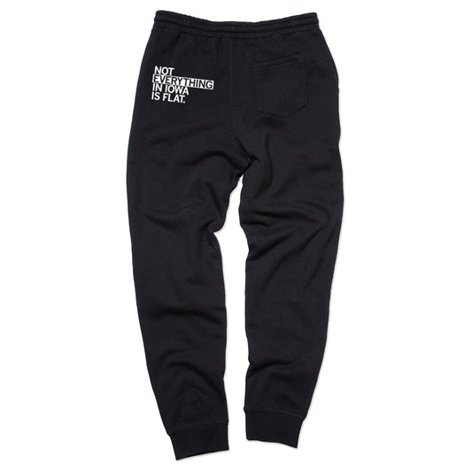 Relaxed fit black sweatpants with white text printed on the back pocket area that reads, NOT EVERYTHING IN IOWA IS FLAT.