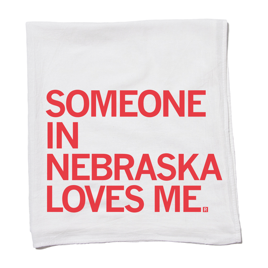 A flour sack towel made of white fabric with the red text, SOMEONE IN NEBRASKA LOVES ME.