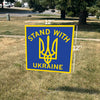 A 12-inch square Stand With Ukraine yard sign featuring a yellow Ukrainian trident symbol on a lawn.