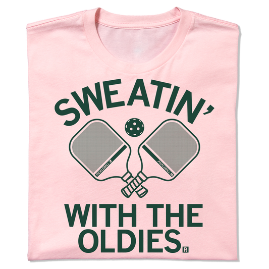A light pink, union-made pickleball t-shirt featuring a green graphic of two paddles, a ball, and the text Sweatin with the Oldies.