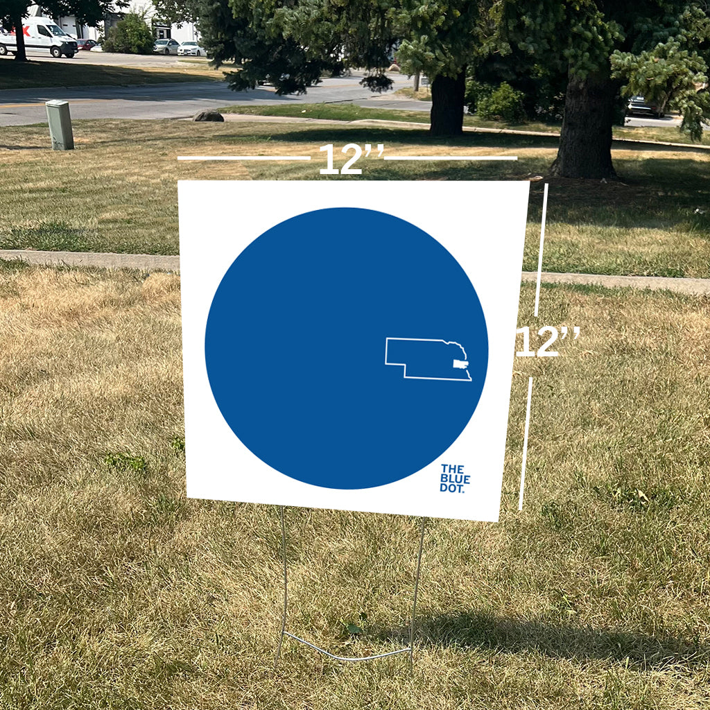 A 12-inch by 12-inch square RAYGUN yard sign featuring a large blue circle with a white outline of Nebraska and the text THE BLUE DOT.