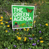 A 12-inch by 12-inch square RAYGUN chloroplast yard sign reading The Green Agenda with a monarch butterfly and floral illustrations, displayed in a patch of dandelions and purple flowers.