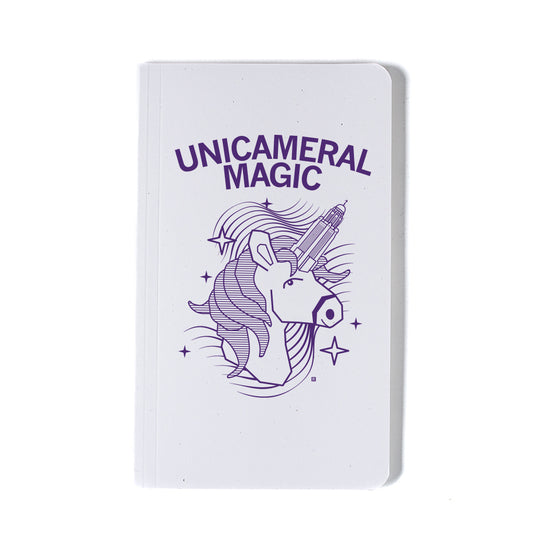 White notebook featuring a dark purple screen print of a unicorn with a camera lens for a horn and the text UNICAMERAL MAGIC.