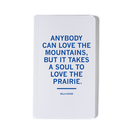 A white notebook featuring the cobalt blue screen print: Anybody can love the mountains, but it takes a soul to love the prairie, a quote attributed to Willa Cather.