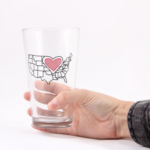 A hand holds a dishwasher-safe Midwest pint glass featuring a black outline of the United States with a red heart icon over the center.