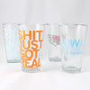 Four clear Iowa pint glasses featuring various slogans and designs, including SHIT JUST GOT REAL in large orange letters printed with soy-based ink.