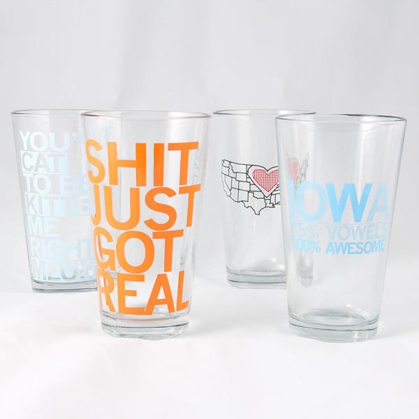 Four clear Iowa pint glasses featuring various slogans and designs, including SHIT JUST GOT REAL in large orange letters printed with soy-based ink.