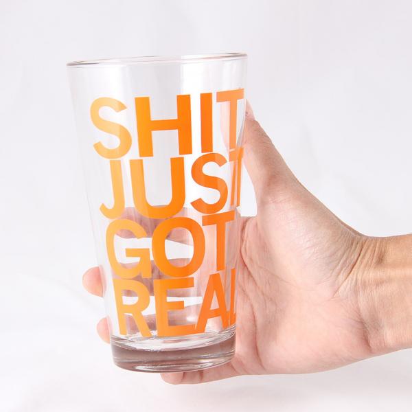 A hand holds a dishwasher-safe pint glass printed with the orange text SHIT JUST GOT REAL.