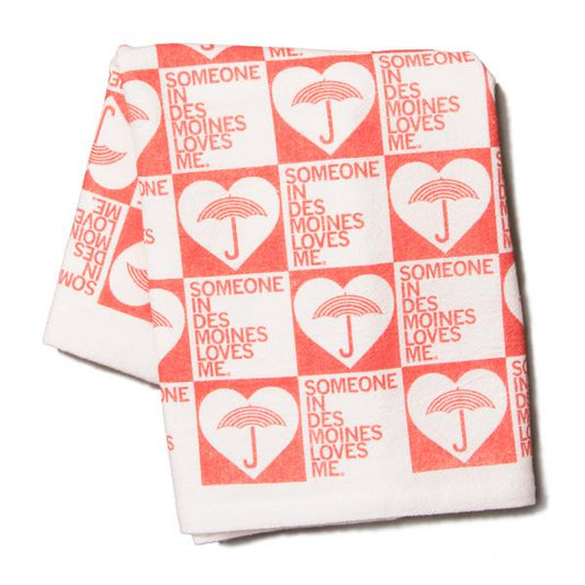 White kitchen towel featuring a repeating red grid pattern, with each square containing a heart with an umbrella inside and the text Someone in Des Moines loves me.